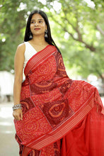 Load image into Gallery viewer, A woman wearing a red and maroon patterned sambalpuri saree with a white sleeveless blouse, standing outdoors against a blurred green background.